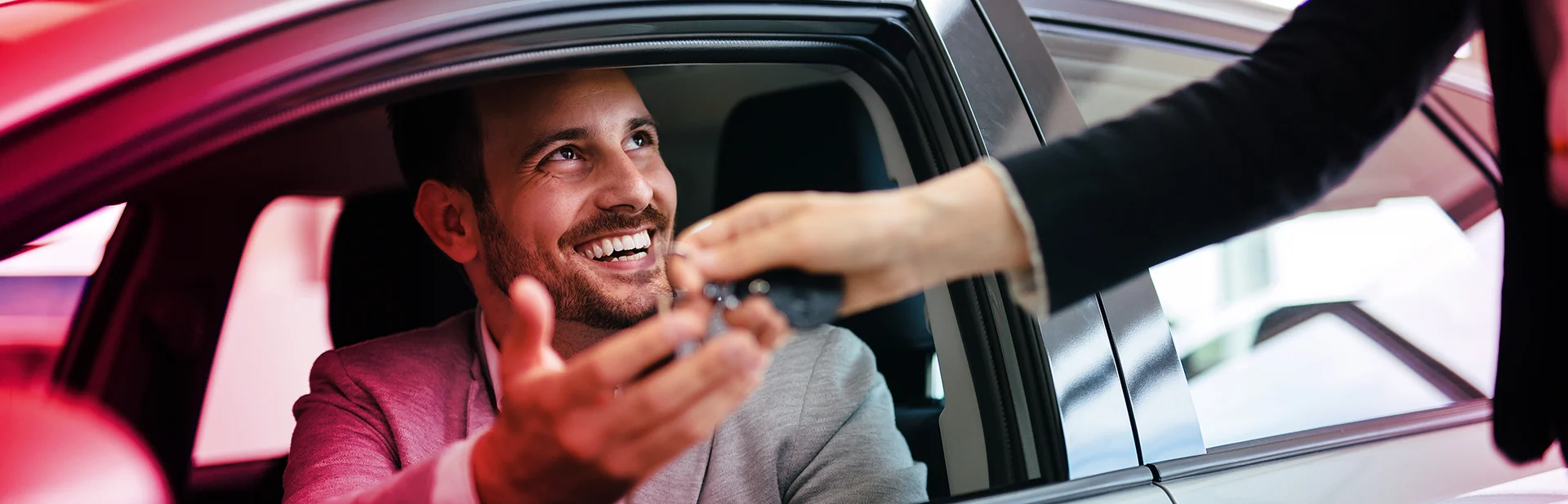 Ein Mann sitzt in einem Auto, Scheibe ist geöffnet und eine Hand einer anderen Pereson überreicht durch das Fenster einen Autoschlüssel